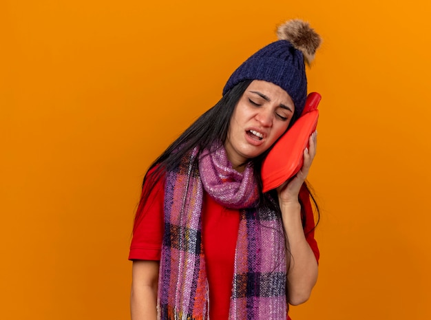 Bolesna młoda kaukaska chora dziewczyna w czapce zimowej i szaliku dotykająca twarzy z torbą na gorącą wodę z zamkniętymi oczami odizolowana na pomarańczowej ścianie z miejscem na kopię