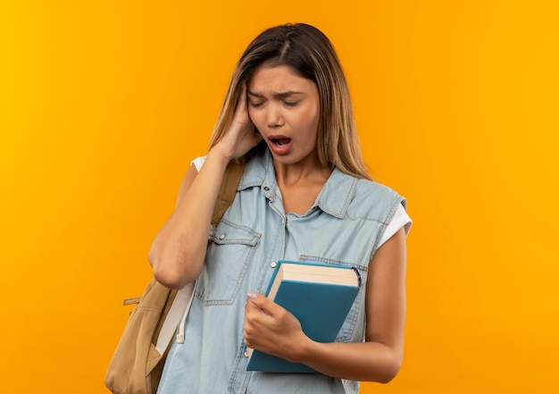 Bolesna młoda ładna studencka dziewczyna nosząca tylną torbę trzymając książkę kładącą rękę na głowie cierpiącą na ból głowy z zamkniętymi oczami odizolowanymi na pomarańczowo z miejscem na kopię