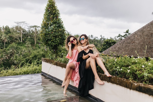 Boso kobieta w czarnej sukni z siostrą w pobliżu basenu. Śmiejąc się modelki w okularach przeciwsłonecznych relaks na Bali w wakacje.