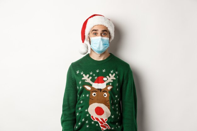 Boże Narodzenie podczas pandemii, koncepcja covid-19. Młody człowiek w santa hat i maska świętuje przyjęcie sylwestrowe ze środkami zapobiegawczymi, białe tło.