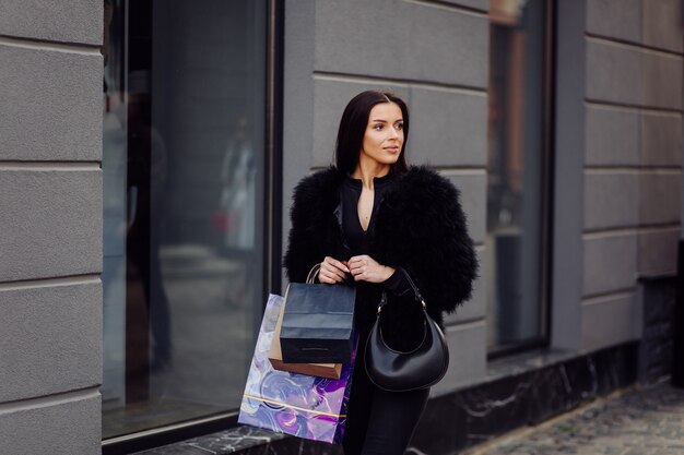 Brązowowłosa kobieta w czarnym stroju trzyma kolorowe, wzorzyste torby na zakupy podczas udanych zakupów. Wychodząc na zewnątrz, cieszy się ciepłem dnia