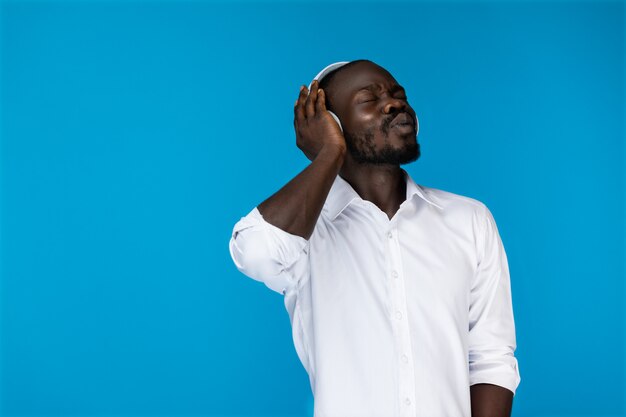 Brodaty afroamerican mężczyzna z zamkniętymi oczami trzyma jedną ręką duże słuchawki w białej koszuli