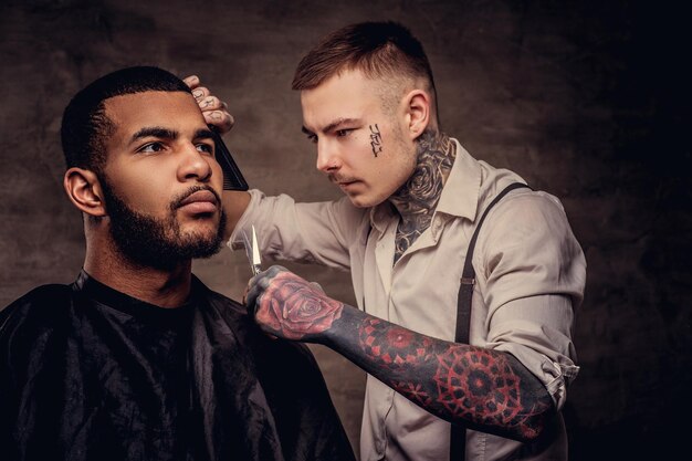 Brodaty Afroamerykański hipster strzyżony przez staromodnego profesjonalnego fryzjera z tatuażem robi strzyżenie.