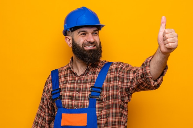 Bezpłatne zdjęcie brodaty budowniczy mężczyzna w mundurze budowlanym i kasku ochronnym, patrząc na bok, uśmiechnięty radośnie szczęśliwy i pozytywny pokazując kciuk w górę