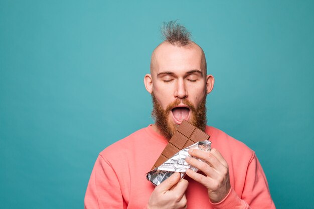 Brodaty Europejczyk w swobodnej brzoskwini na białym tle, ciesząc się kawałkiem czekolady z zamkniętymi oczami