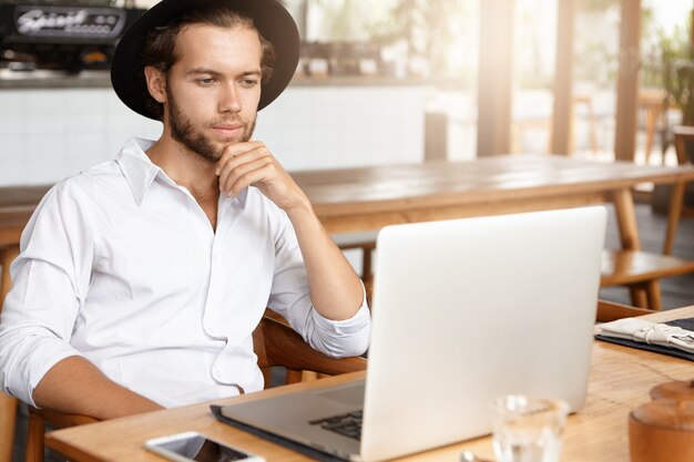 Brodaty freelancer łączący się z siecią bezprzewodową przez laptopa. Przemyślany mężczyzna pracuje na notebooku, siedząc przy drewnianym stole w nowoczesnym wnętrzu kawiarni. Student czytanie książki w kawiarni