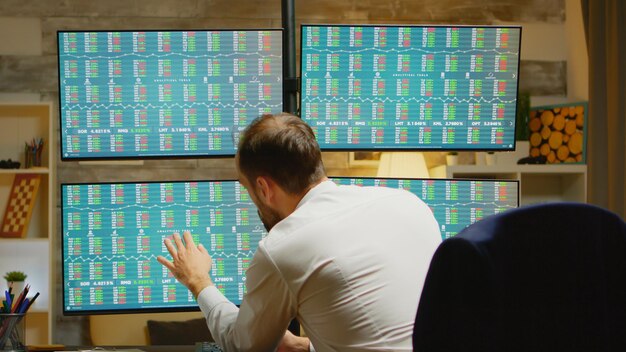 Brodaty handlarz w swoim domowym biurze w garniturze sprawdzający giełdę na czterech ekranach