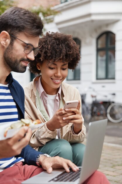 Brodaty hipster przegląda witrynę sieciową, je fast foody. Afroamerykańska dziewczyna używa telefonu komórkowego