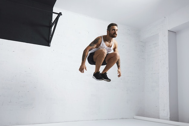 Brodaty i wytatuowany młody sportowiec pokazuje ruchy kalisteniczne, skacze wysoko w powietrzu obok czarnego drążka, ubrany w koszulkę z pustym czołgiem, odizolowany w białym pokoju centrum fitness