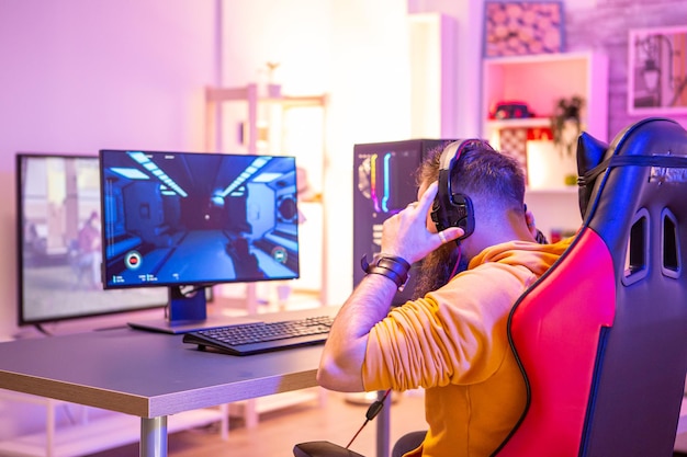 Brodaty mężczyzna grający w gry wideo online na swoim komputerze i rozmawiający z innymi graczami. W pokoju kolorowe neony.