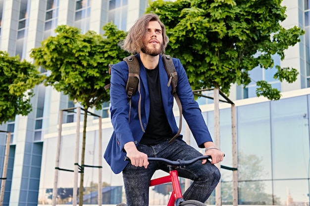 Brodaty mężczyzna hipster z plecakiem siedzi na czerwonym rowerze stacjonarnym w parku.