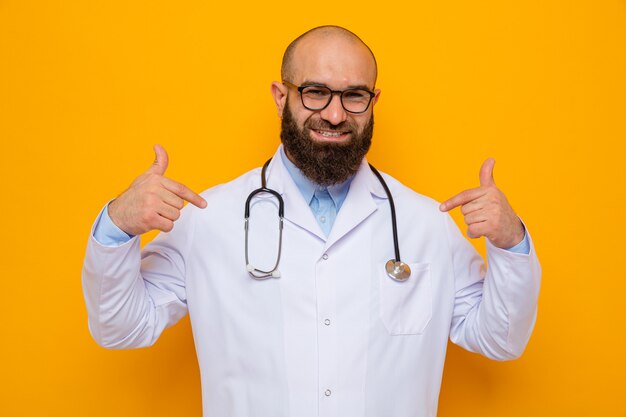Brodaty mężczyzna lekarz w białym fartuchu ze stetoskopem na szyi w okularach wyglądający uśmiechnięty pewnie wskazujący na siebie