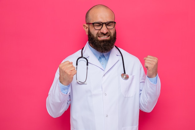 Brodaty mężczyzna lekarz w białym fartuchu ze stetoskopem na szyi w okularach zaciskając pięści sfrustrowany i zły stojąc na różowym tle