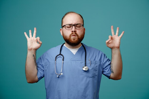 Brodaty mężczyzna lekarz w mundurze ze stetoskopem na szyi w okularach medytuje wykonując gest medytacji palcami spokojny i zrelaksowany stojąc na niebieskim tle