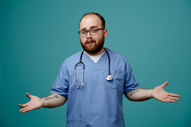 Brodaty mężczyzna lekarz w mundurze ze stetoskopem na szyi w okularach, patrząc na kamerę zdezorientowany rozkładając ręce na boki, nie mając odpowiedzi, stojąc na niebieskim tle