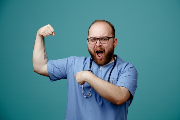 Bezpłatne zdjęcie brodaty mężczyzna lekarz w mundurze ze stetoskopem na szyi w okularach wyglądający na podekscytowanego i silną unoszącą pięść pokazującą bicepsy stojące na niebieskim tle