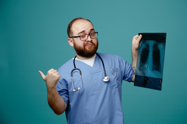 Brodaty mężczyzna lekarz w mundurze ze stetoskopem wokół szyi w okularach trzymających xray płuc patrząc na kamerę uśmiechający się pewnie pokazując kciuk do góry stojący na niebieskim tle