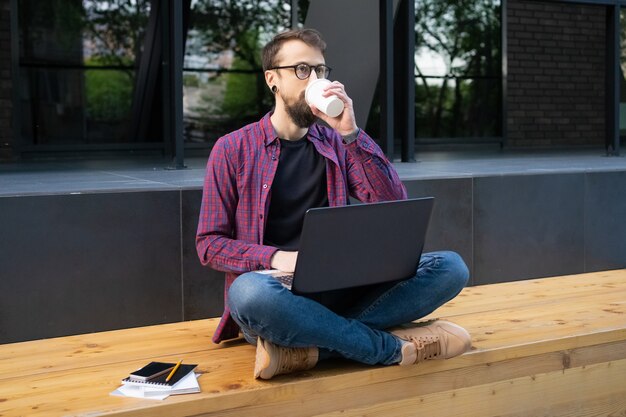 Brodaty mężczyzna siedzący ze skrzyżowanymi nogami na drewnianej ławce z laptopem