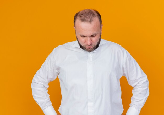 Brodaty mężczyzna ubrany w białą koszulę, patrząc w dół z poważną twarzą stojącą na pomarańczowej ścianie