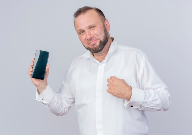 Brodaty Mężczyzna Ubrany W Białą Koszulę Pokazuje Smartfon Uśmiechnięty Pewny Siebie Stojący Na Białej ścianie