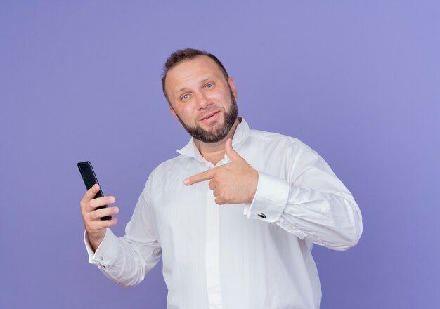 Brodaty mężczyzna ubrany w białą koszulę, trzymając smartfon, wskazując palcem na niego uśmiechnięty, stojąc na niebieskiej ścianie