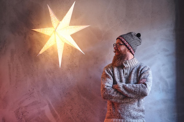 Brodaty mężczyzna ubrany w ciepły sweterek i czapkę na ścianę z ozdobną lampą w gwiazdkę.