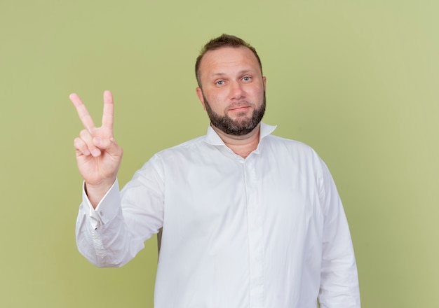 Brodaty mężczyzna w białej koszuli pokazuje palcami numer dwa i wskazuje w górę z poważną twarzą stojącą nad jasną ścianą