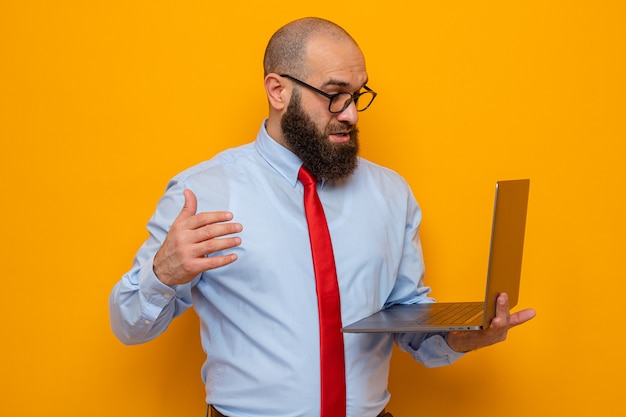 Brodaty mężczyzna w czerwonym krawacie i niebieskiej koszuli w okularach, trzymający laptopa, patrząc na niego zdumiony i zaskoczony, stojąc na pomarańczowym tle