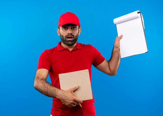 Brodaty mężczyzna w czerwonym mundurze i czapce trzymającej pudełko pokazujące schowek patrząc zaskoczony stojąc nad niebieską ścianą
