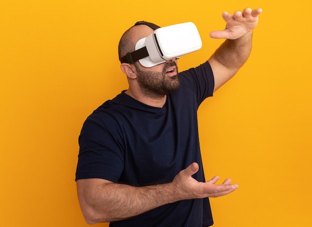Brodaty mężczyzna w granatowej koszulce w okularach wirtualnej rzeczywistości, gestykuluje rękami stojącymi na pomarańczowej ścianie