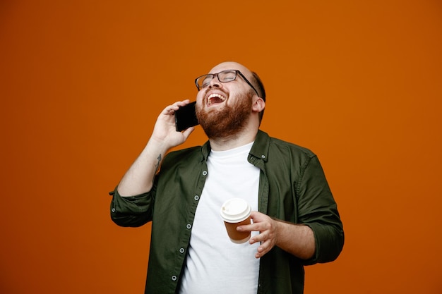 Brodaty mężczyzna w zwykłych ubraniach w okularach rozmawia przez telefon komórkowy, trzymając kubek kawy szczęśliwy i podekscytowany, śmiejąc się stojąc na pomarańczowym tle
