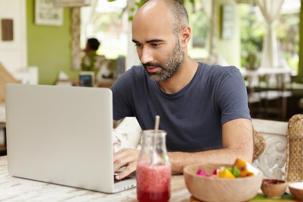 Brodaty samozatrudniony mężczyzna w średnim wieku siedzi w kawiarni przed zwykłym laptopem i patrzy na ekran z poważną i skoncentrowaną miną podczas pracy zdalnej nad swoim projektem, korzystając z bezpłatnego Wi-Fi