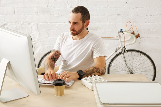 Brodaty wytatuowany freelancer w pustej białej koszulce pracuje na swoim komputerze w domu przed murem i zaparkowanym rowerem vintage, czas letni