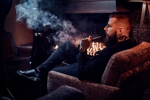 Brodaty zrelaksowany mężczyzna siedzi na fotelu i pali fajkę wodną przy kominku.