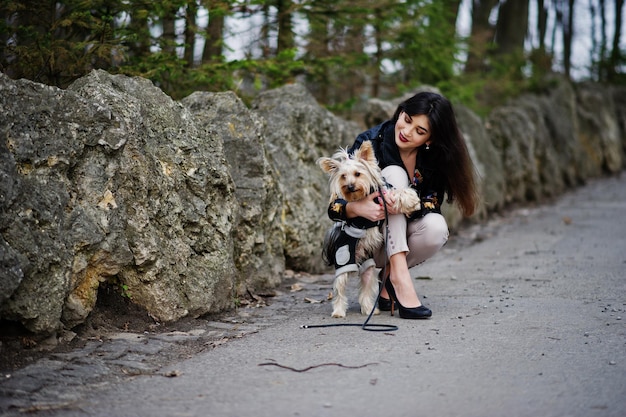 Bezpłatne zdjęcie brunetka cygańska dziewczyna z psem yorkshire terrierem na tle kamieni w parku model nosi skórzaną kurtkę i koszulkę z ozdobnymi spodniami i butami na wysokich obcasach