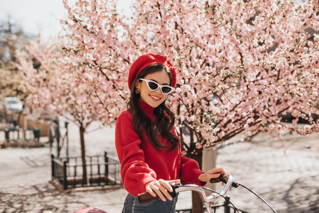 Brunetka dziewczyna w czerwonym kapeluszu i swetrze, pozowanie na tle sakury. Urocza kobieta to stylowe okulary przeciwsłoneczne, uśmiechnięta i jeżdżąca na rowerze