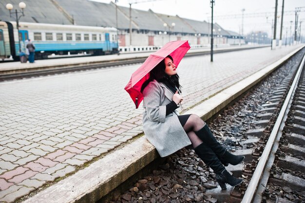Brunetka dziewczyna w szarym płaszczu z czerwonym parasolem na stacji kolejowej