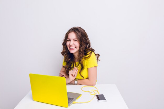 Brunetka dziewczyna z kręconymi włosami śmieje się przed laptopem w żółtej obudowie.