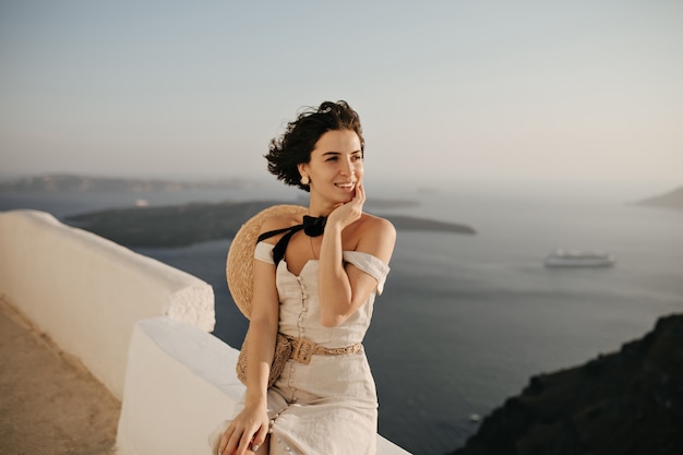 Brunetka krótkowłosa kobieta w eleganckiej beżowej sukience i wioślarzu cieszy się pięknym widokiem na morze