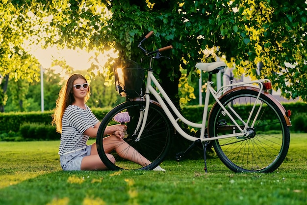 Brunetka siedzi na zielonym trawniku z rowerem i trzyma bukiet kwiatów w parku.