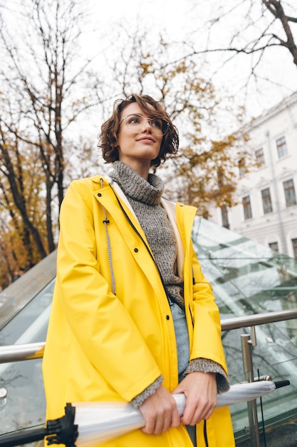 Brunetki kobieta bada ostrzyżenie jest ubranym szkła i żółtego deszczowiec bada punkty zwrotnych w turystycznym miejscu