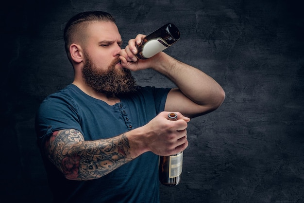 Brutalny brodaty mężczyzna z wytatuowanym ramieniem pije piwo z butelki.
