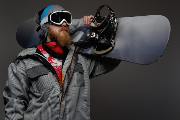 Bezpłatny plik Zdjęcie brutalny mężczyzna z rudą brodą w pełnym ekwipunku, trzymający na ramieniu deskę snowboardową, odizolowany na ciemnym tle.
