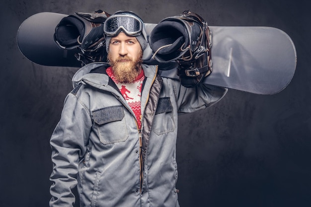 Bezpłatne zdjęcie brutalny rudowłosy snowboardzista z brodą w zimowej czapce i okularach ochronnych, ubrany w snowboardowy płaszcz, trzyma na ramieniu deskę snowboardową w studio. na białym tle na szarym tle.