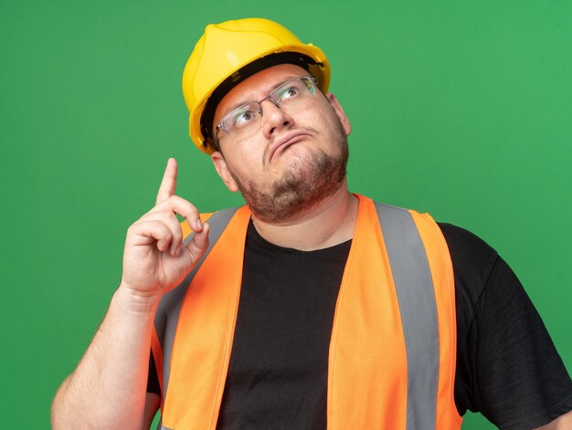 Budowniczy człowiek w kamizelce budowlanej i kasku ochronnym patrzący w górę zdziwiony, wskazujący palec wskazujący w górę, stojący nad zielenią