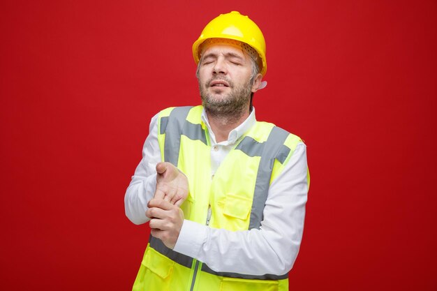 Budowniczy człowiek w mundurze budowlanym w kasku ochronnym wygląda na zmęczonego i przepracowanego, rozciągając ręce stojąc na czerwonym tle