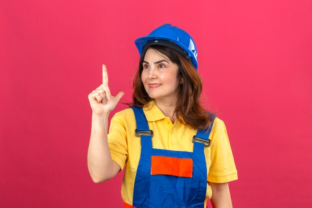 Bezpłatne zdjęcie budowniczy kobieta ubrana w mundur budowlany i kask ochronny, patrząc na bok, wskazując palcem w górę, uśmiechając się, mając nowy pomysł stojący nad izolowaną różową ścianą