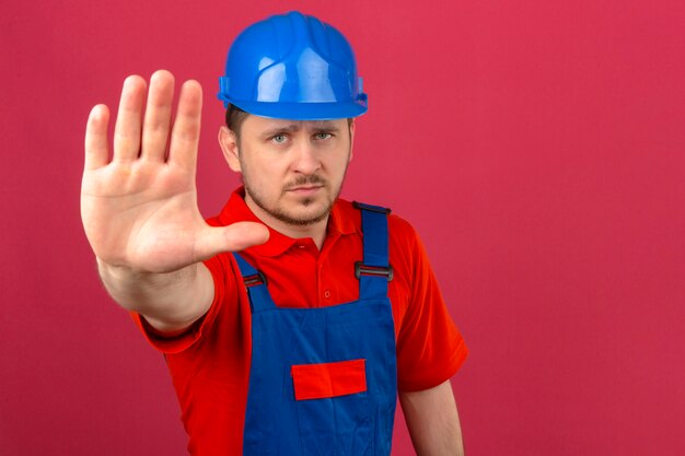 Budowniczy mężczyzna ubrany w mundur budowlany i hełm ochronny stojący z otwartą ręką robi znak stop z poważnym i pewnym siebie gestem obrony na izolowanej różowej ścianie