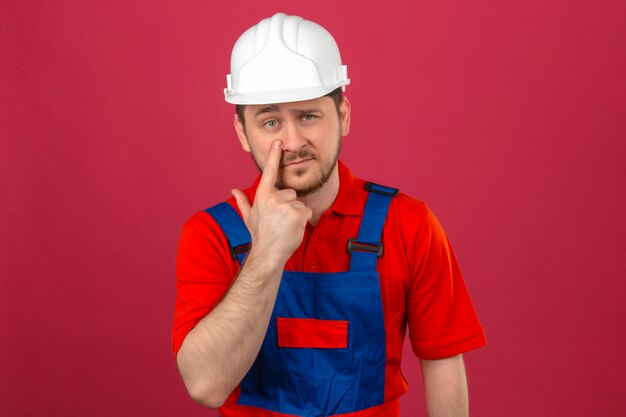 Budowniczy mężczyzna ubrany w mundur budowlany i hełm ochronny, wskazując na oko, patrząc, jak gestykulujesz podejrzaną miną stojącą nad izolowaną różową ścianą