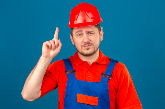 Budowniczy mężczyzna ubrany w mundur budowlany i hełm ochronny, wskazując palcem w górę, uśmiechnięty pewnie, że ma świetny pomysł na odizolowaną niebieską ścianę